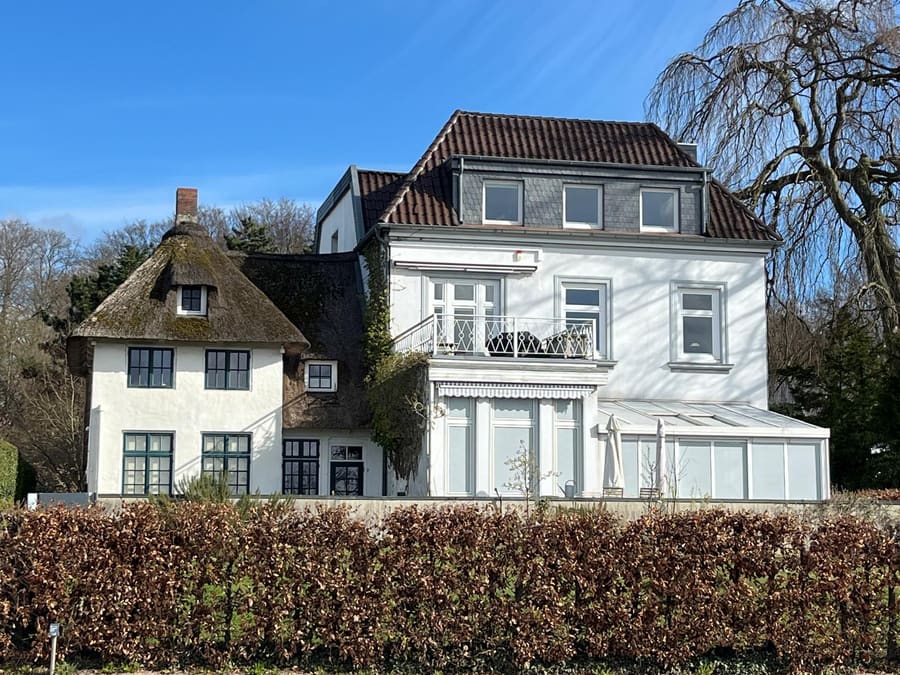 Reetgedecktes Haus neben einer weißen Villa mit Wintergarten hinter einer Buchenhecke