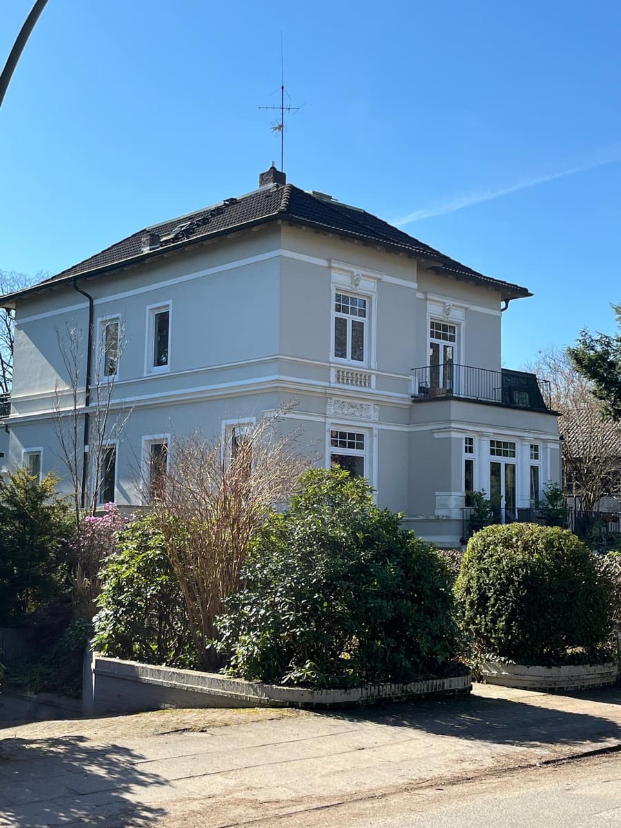 Hellgraue Villa mit Walmdach und Giebel hinter blühenden Sträuchern